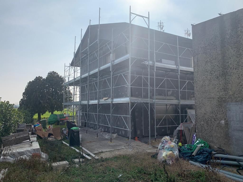 Un'impalcatura circonda un edificio in costruzione, situato accanto a un sentiero sterrato e ad alcuni alberi.