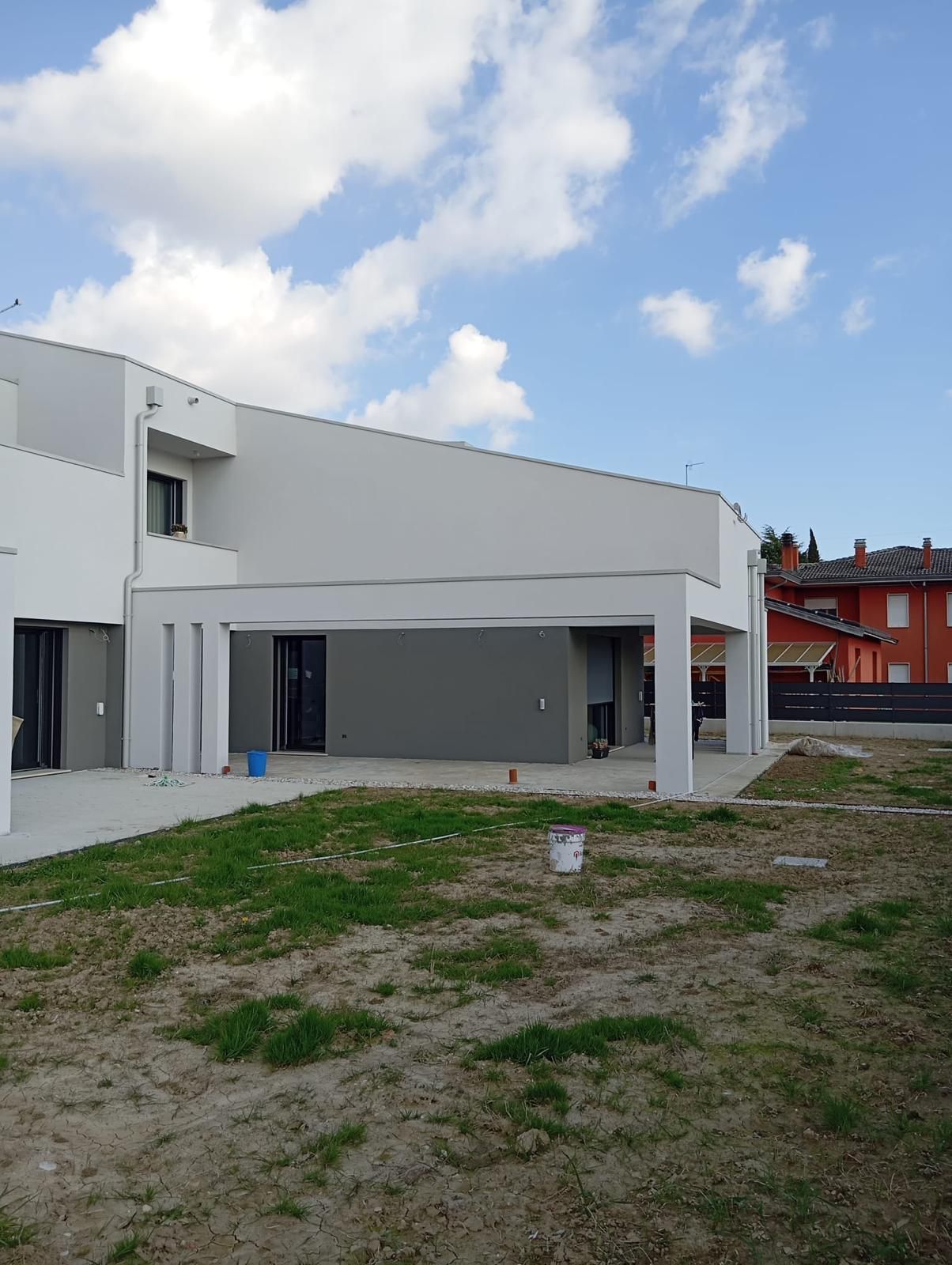 Esterno di una moderna casa bianca con dettagli grigi, giardino non rifinito e cielo azzurro con nuvole.