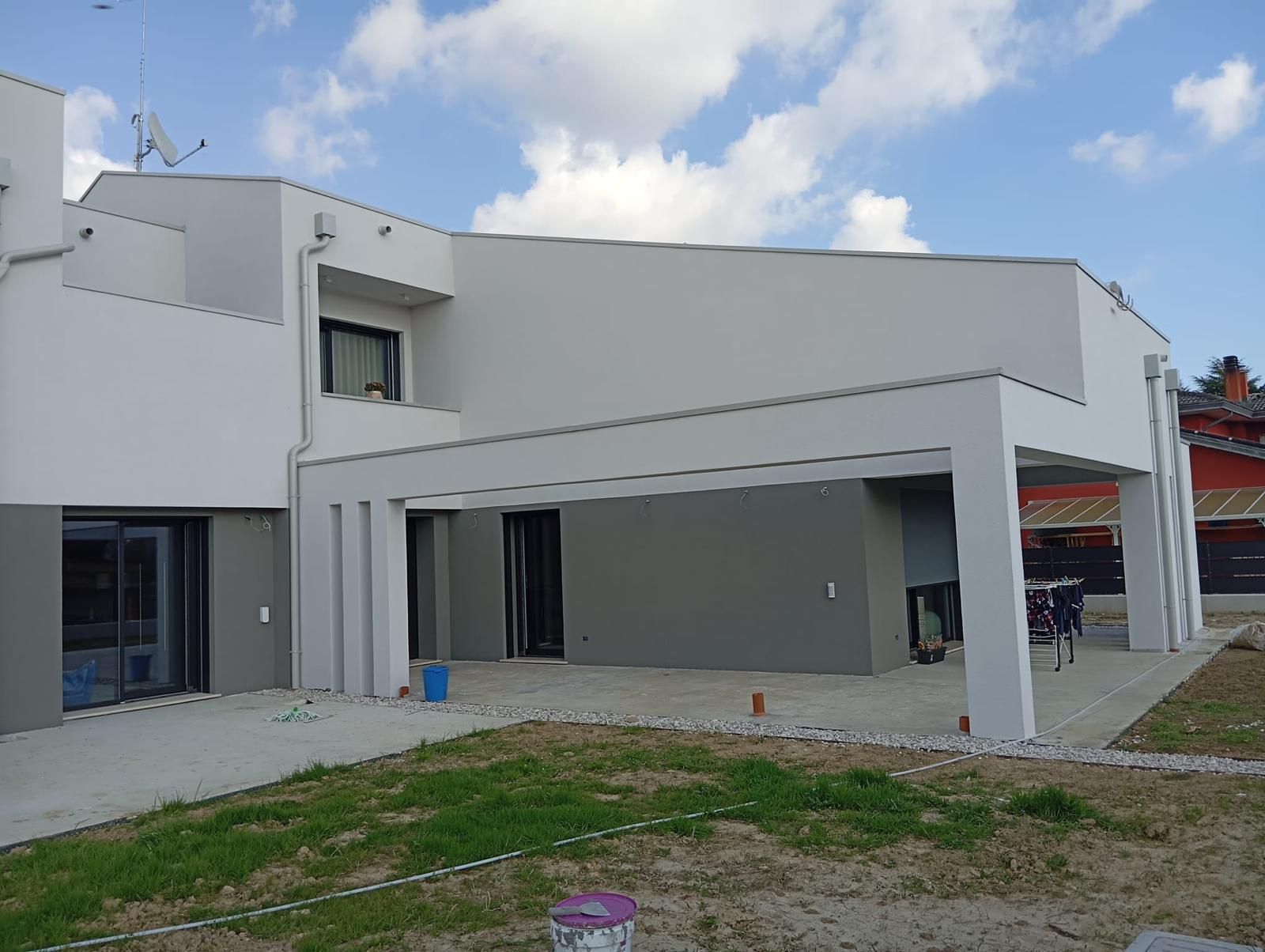 Casa moderna bianca con tetto piano e patio coperto, vista dal giardino sotto un cielo nuvoloso.