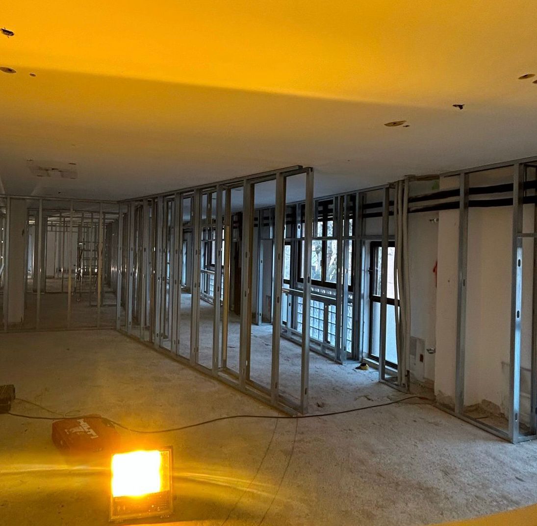Interno parzialmente intelaiato in fase di ristrutturazione, con travi a vista e una luminosa lampada da lavoro sul pavimento.