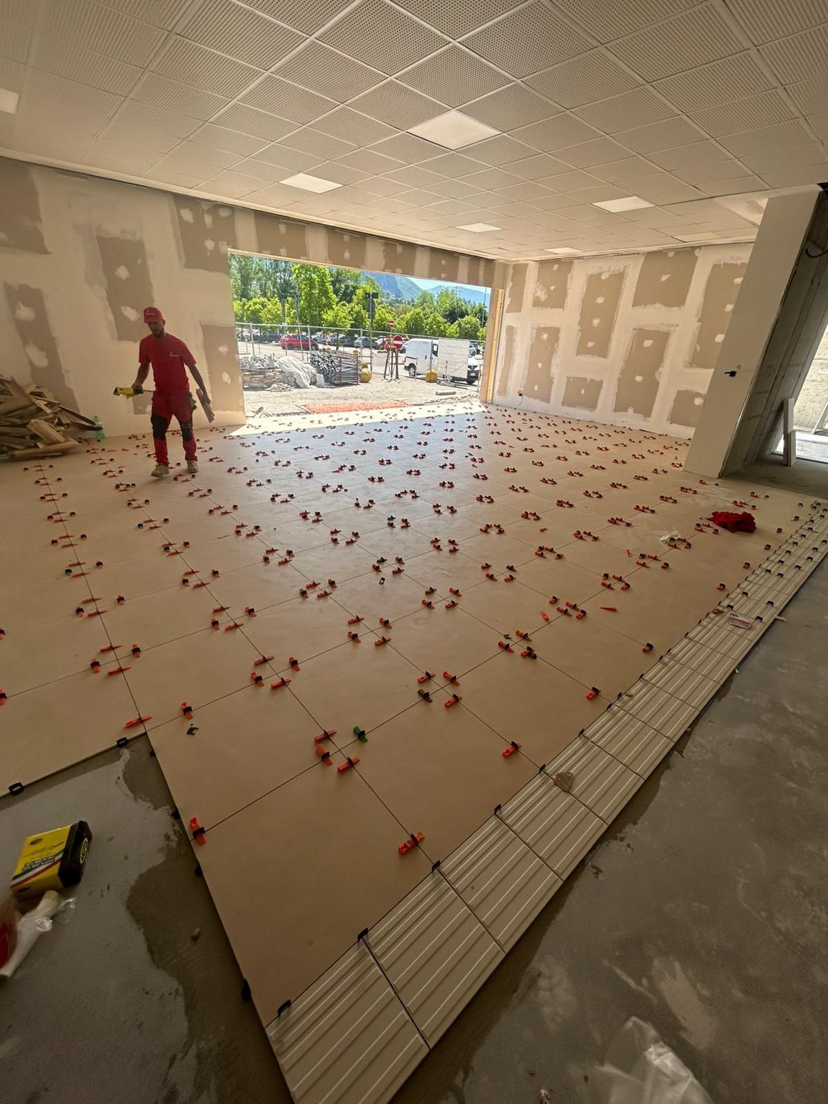 Stanza non finita con pavimento piastrellato, materiali edili e un operaio vicino all'ingresso aperto.