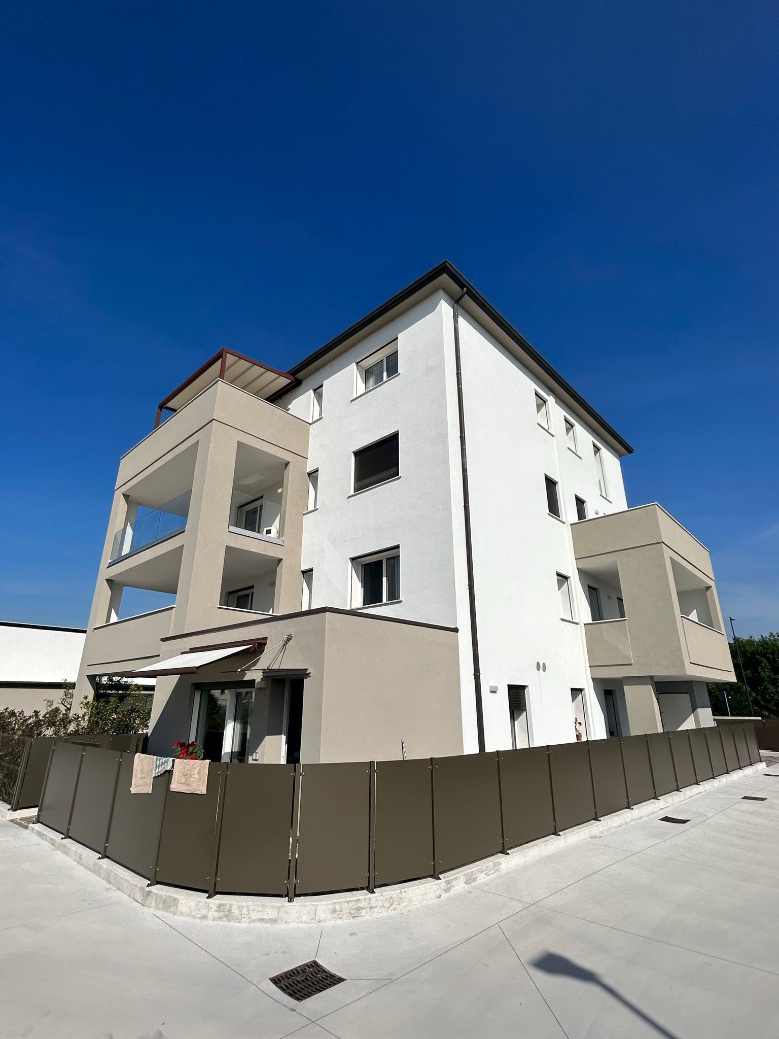 Moderno condominio con facciate bianche e beige dietro un marciapiede recintato sotto un cielo azzurro limpido.
