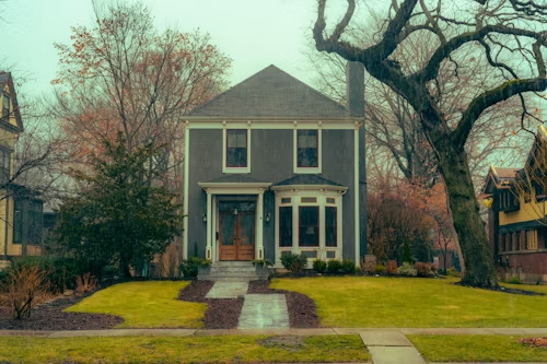 Gray house with bay window, pathway, chimney, and trees on a cloudy day.