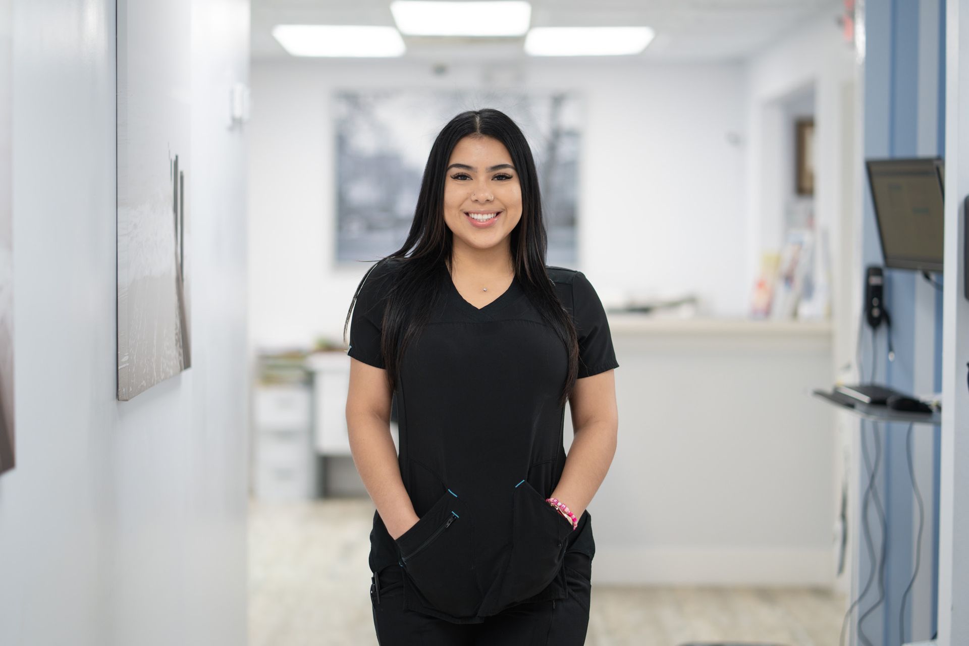 A woman in a black scrub is standing in a hallway with her hands in her pockets and smiling.
