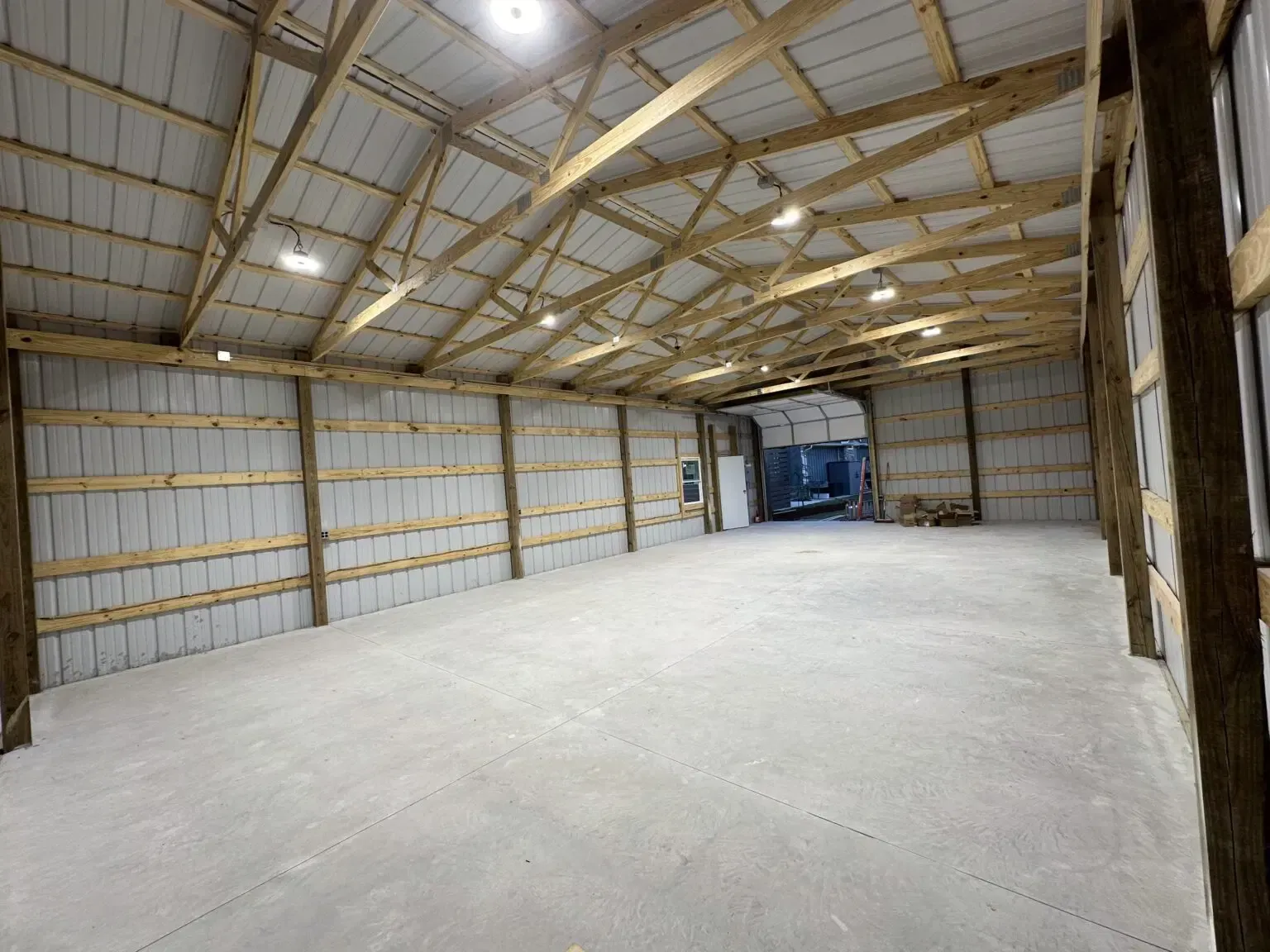 A wide, empty pole barn with a concrete floor, wooden structural beams, white metal walls, and overhead lighting.