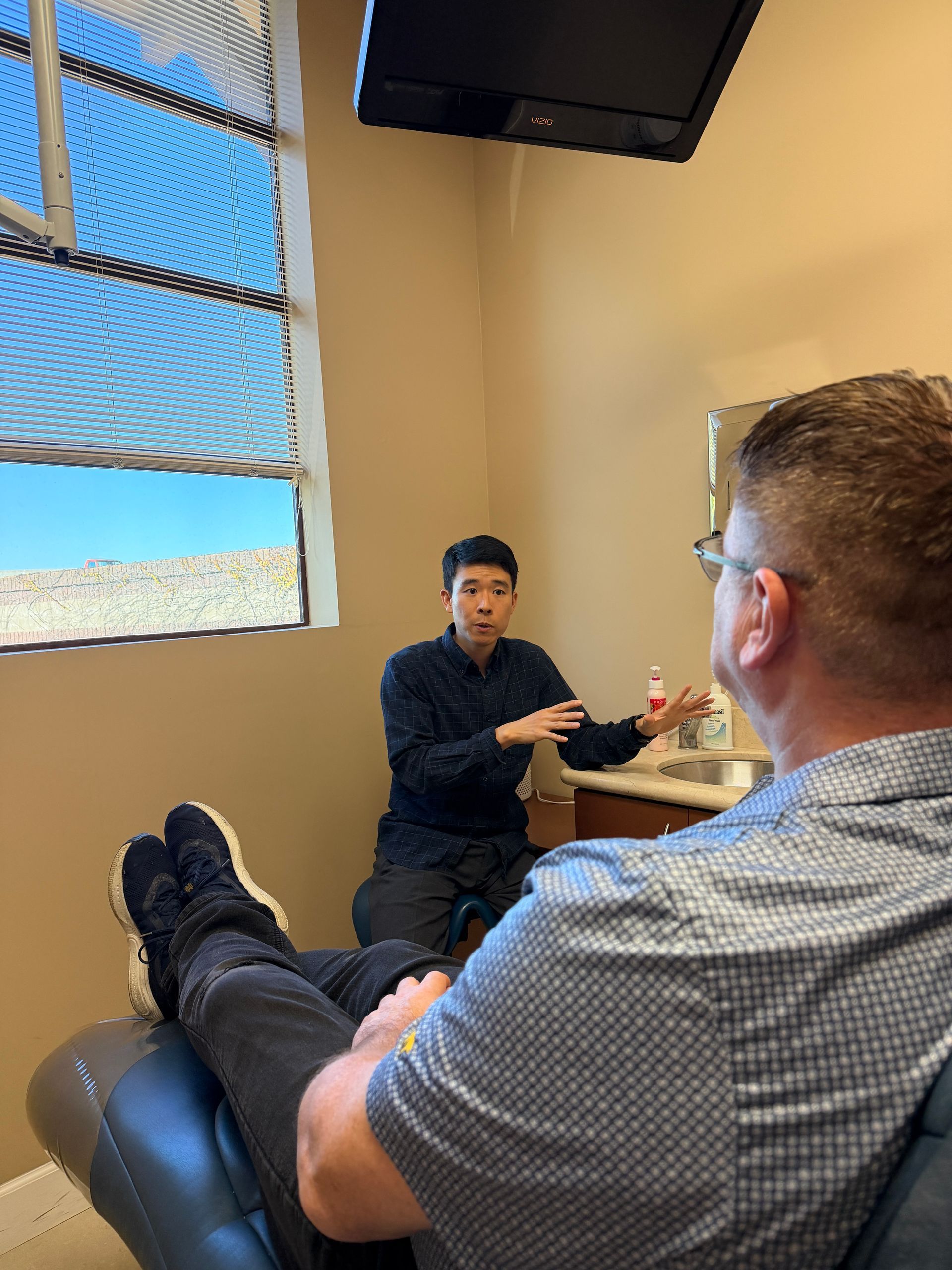 Dentist in blue scrubs consulting a smiling patient holding a tablet in a bright clinic room