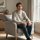 Man sitting in a light gray armchair in a bright living room, wearing a cream sweater and jeans.