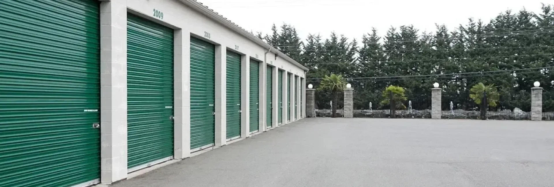 Row of green storage units beside a wide empty paved lot, with trees in the background