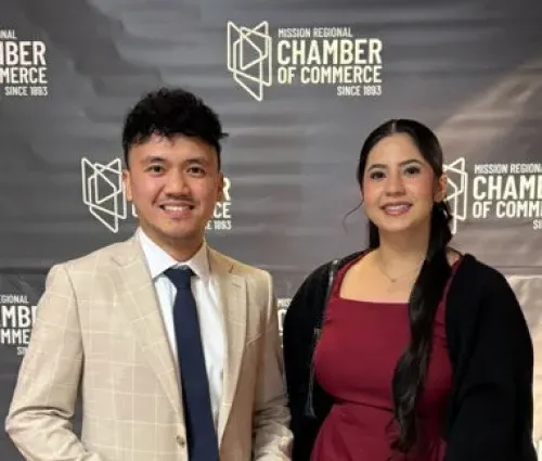 Two people smiling in front of a Chamber of Commerce backdrop, wearing business attire.
