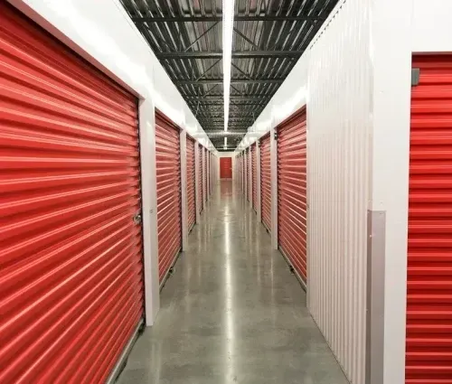 Narrow storage corridor with rows of red roll-up unit doors on both sides, lit by overhead lights