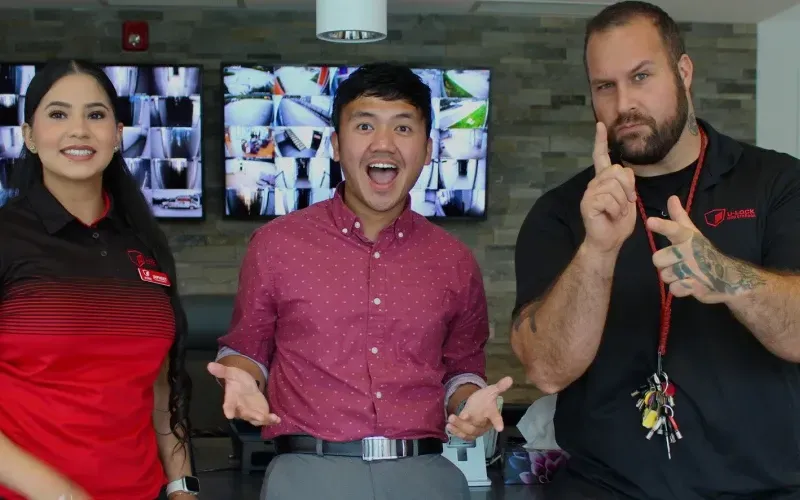 Three people standing indoors, smiling and gesturing in front of wall-mounted screens.