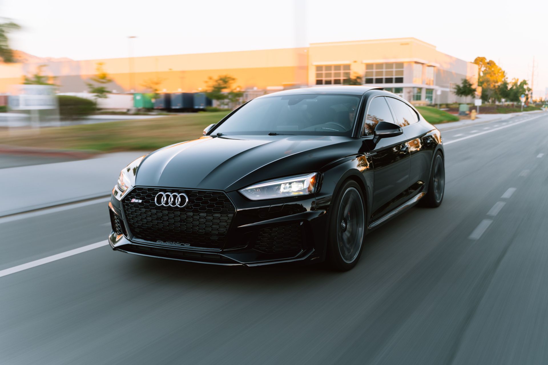 Black Audi sports car driving on a road at dusk, with motion blur and a modern building in the background