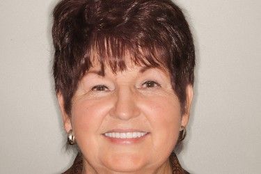 Woman with short brown hair, smiling, showing white teeth, wearing earrings, in front of a neutral background.