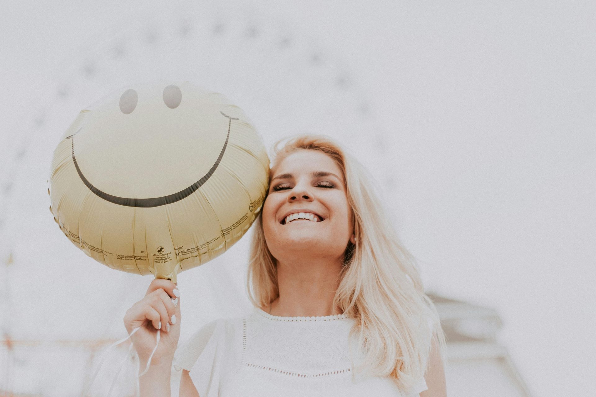 Blonde woman smiling, holding a yellow smiley face balloon. Ferris wheel in background.