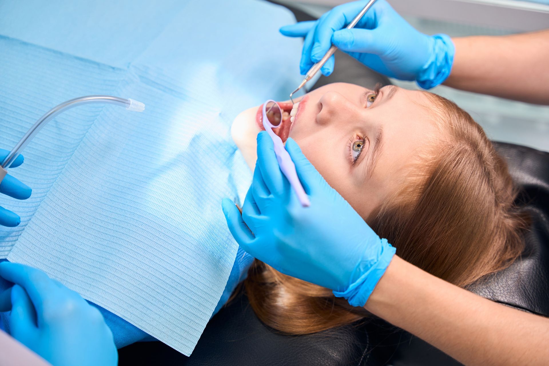 A young girl lies in a dentist's chair with her mouth open, being examined by a dentist wearing blue gloves.