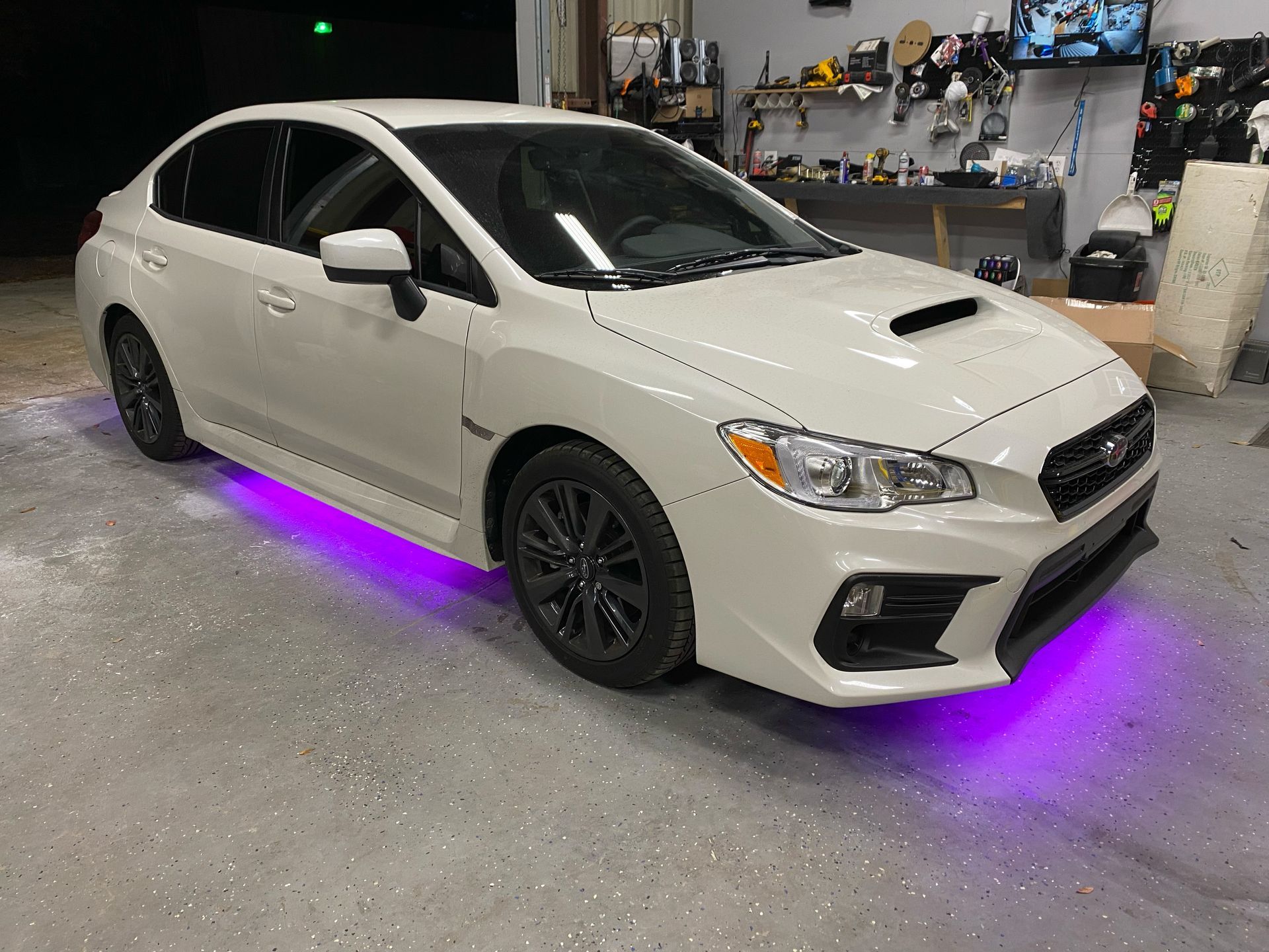 A white subaru wrx with purple lights underneath it is parked in a garage.