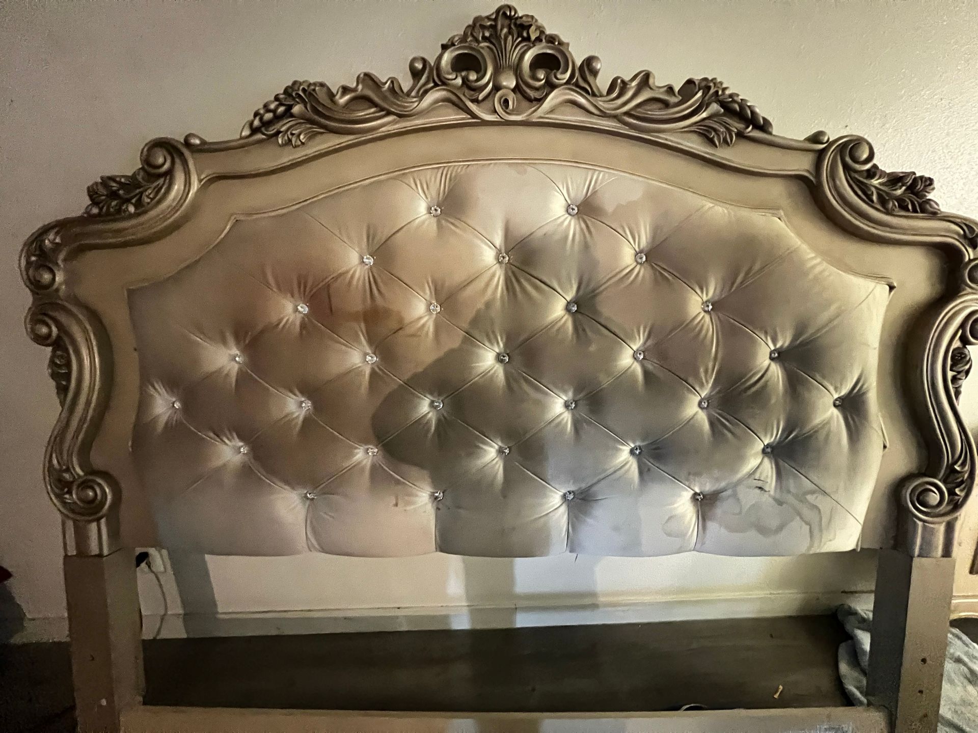 Ornate, tufted headboard, possibly a bed, with light-colored fabric and gold-colored trim.