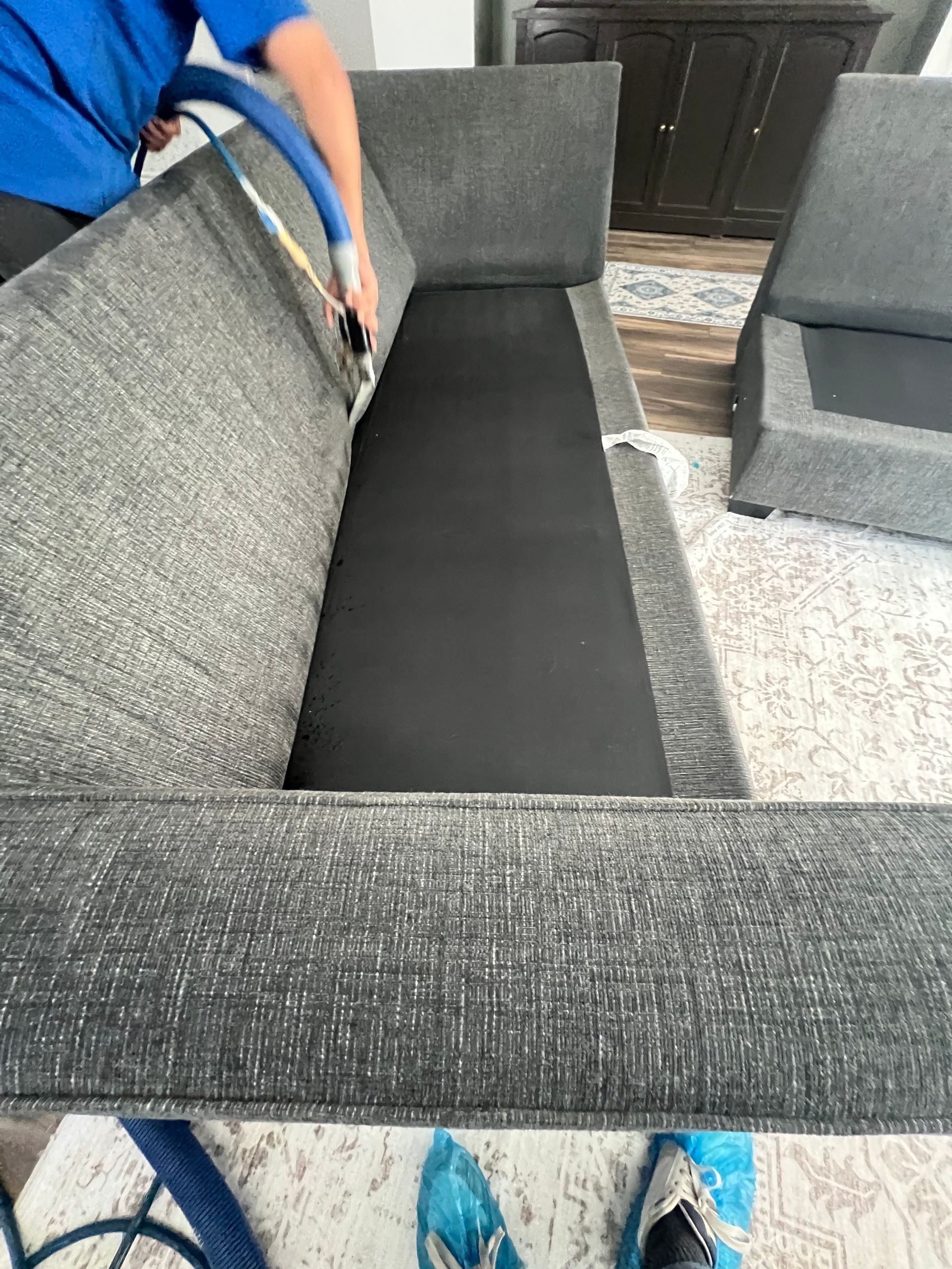 Person vacuuming a gray couch with a black interior, next to an ottoman, on a patterned rug.