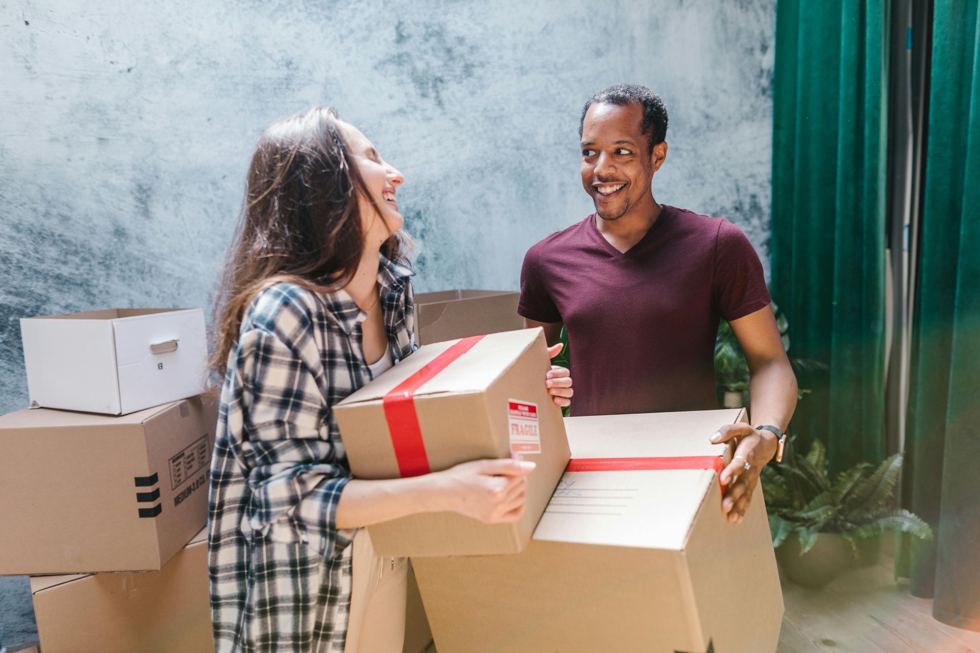 Couple moving boxes, smiling. Boxes piled in room with green curtains.