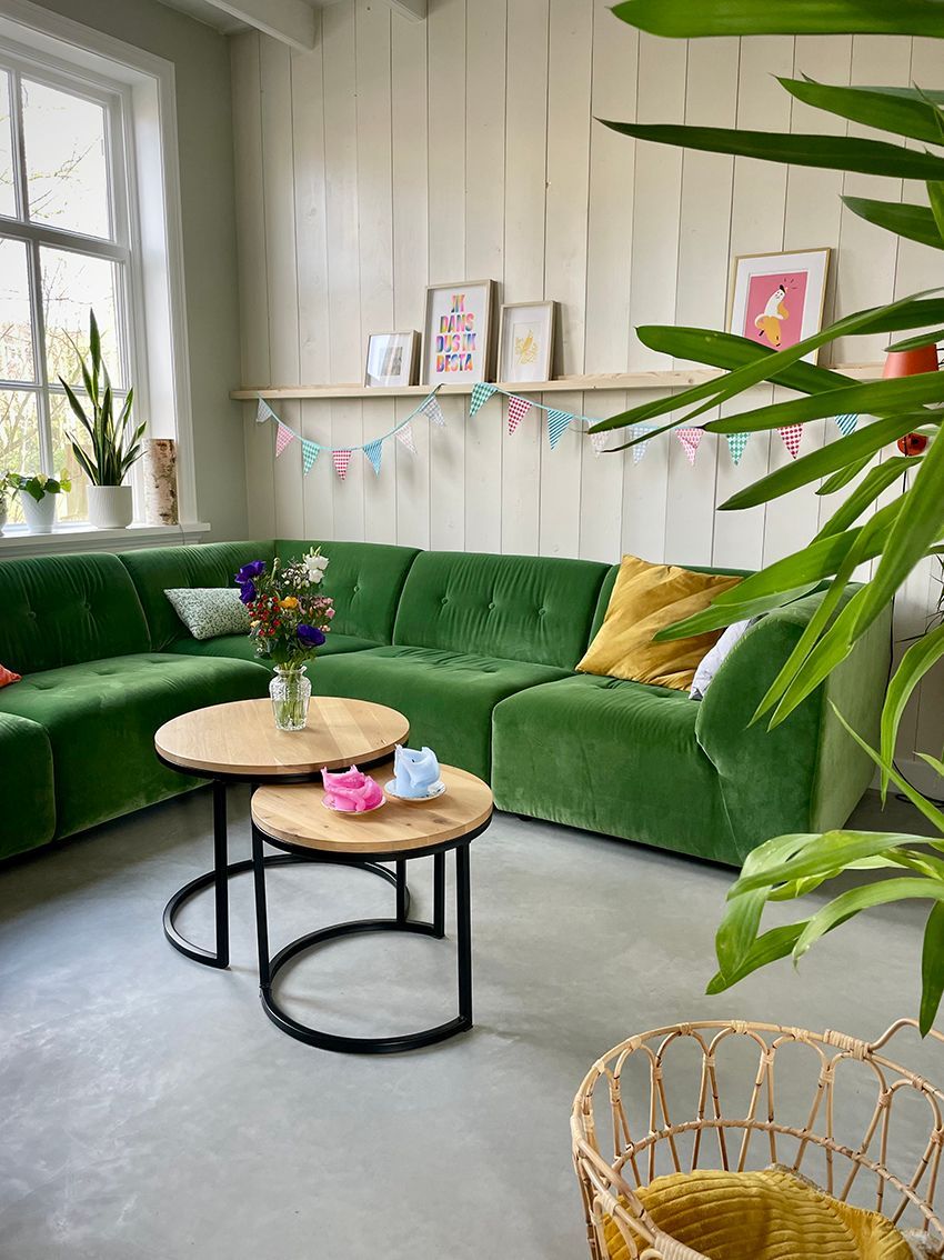 Groene hoekbank in een lichte woonkamer met ronde houten tafels, planken en groene planten.