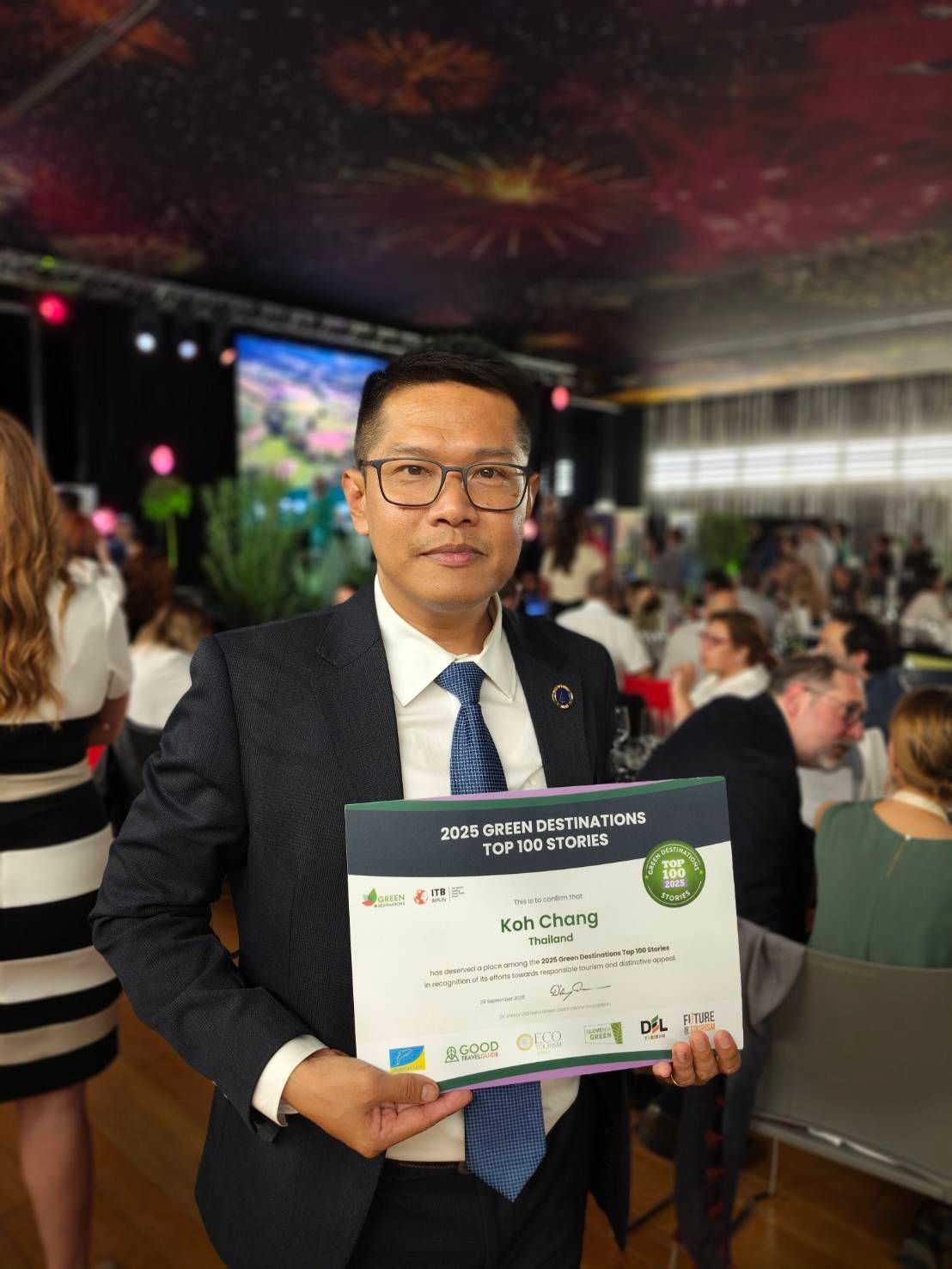 Man in suit holding certificate at an event, surrounded by people.