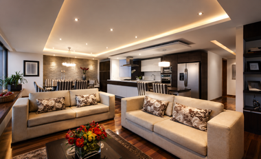 Modern open-concept living room with beige sofas, recessed lighting, and a kitchen in the background