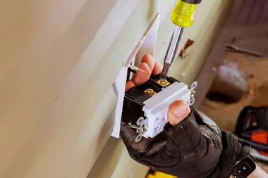 Person wiring an electrical outlet with a screwdriver and voltage tester in a wall opening