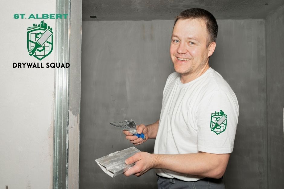 A drywall repair contractor smiles while holding a putty knife and sanding block at a commercial construction site, smoothing drywall in an unfinished room near 255 St, and Hwy 16A, Acheson, AB.