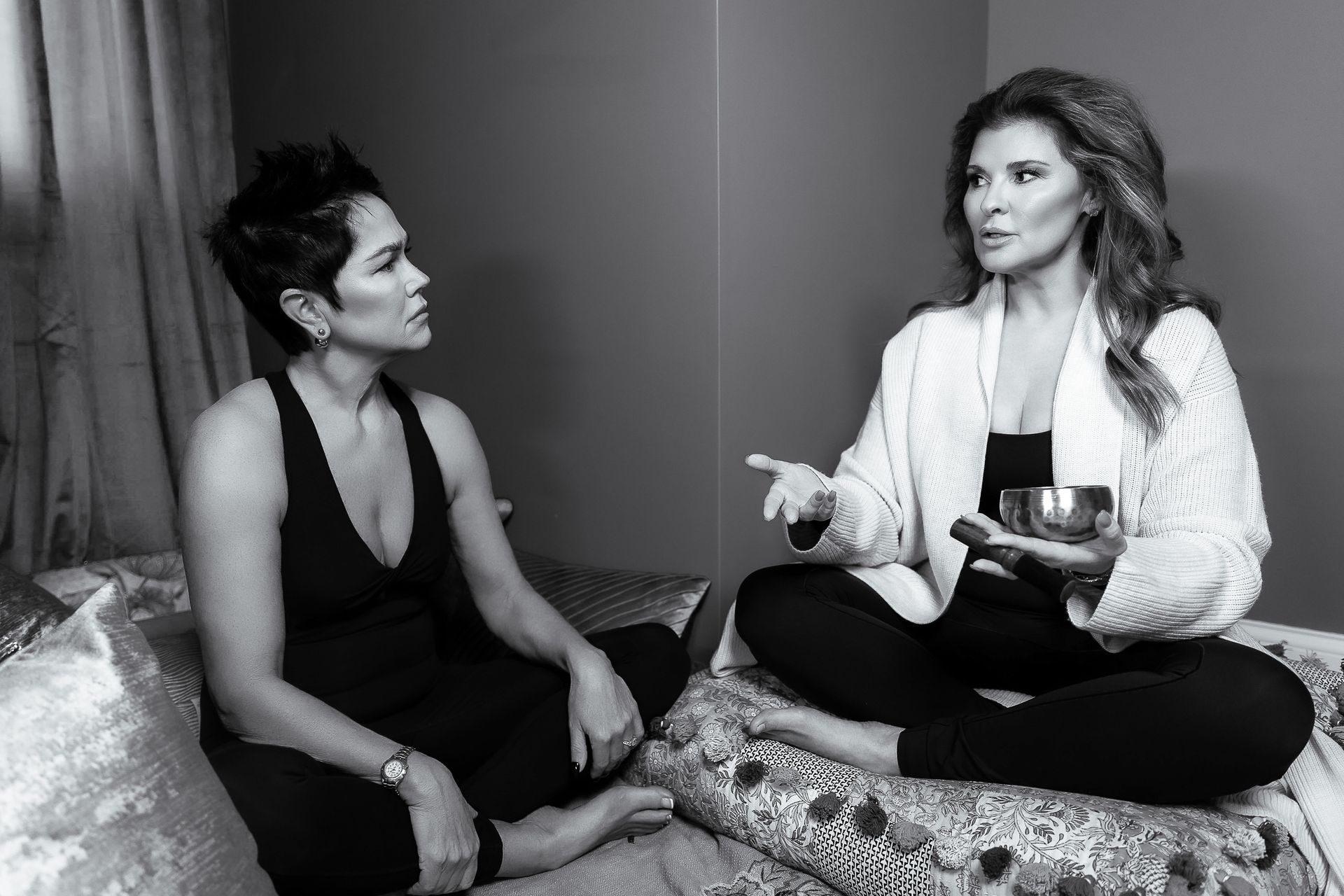 Two women seated on a bed, one holding a singing bowl, talking. Black and white.