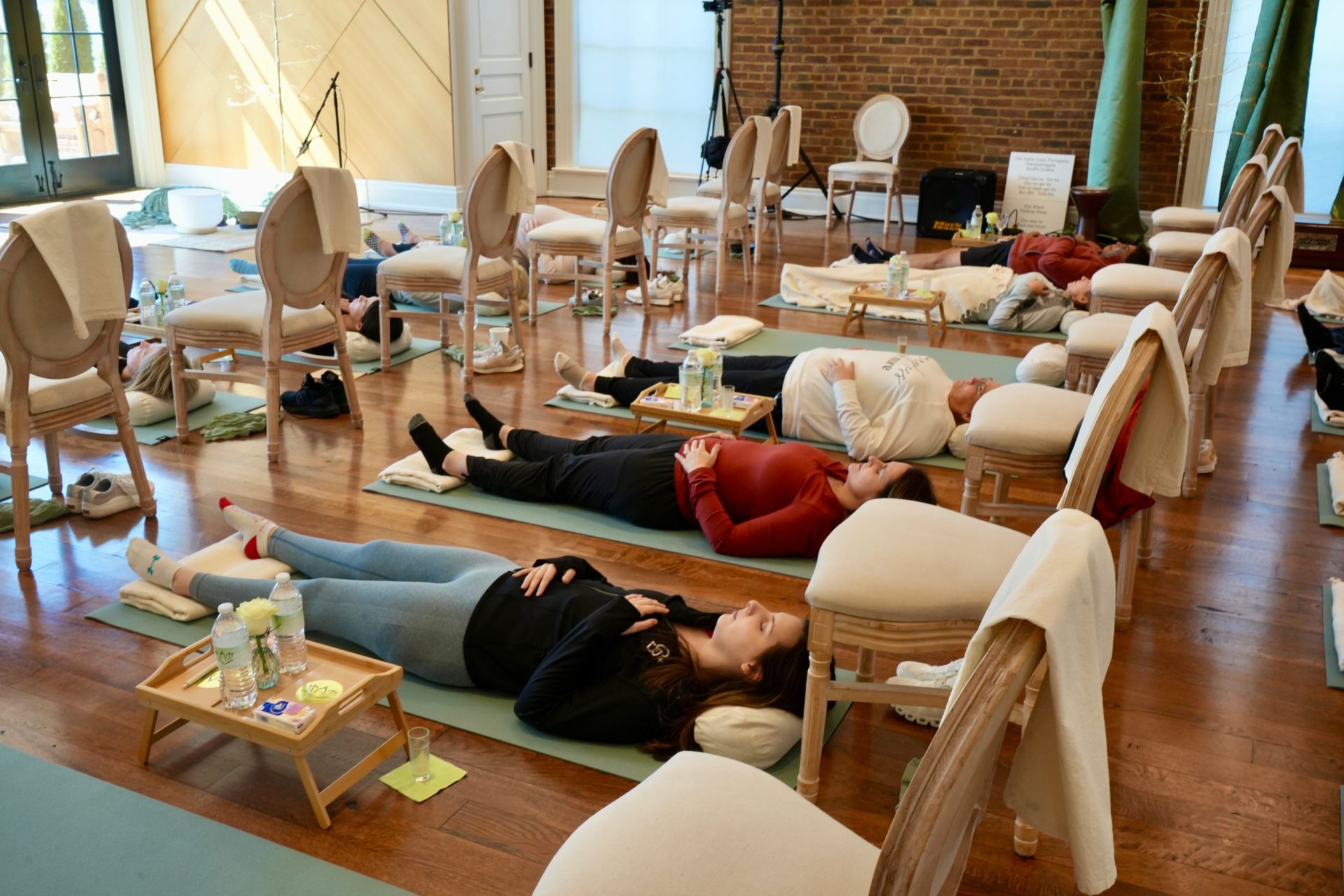 People lying on yoga mats in a room. Chairs with towels are positioned around the mats.