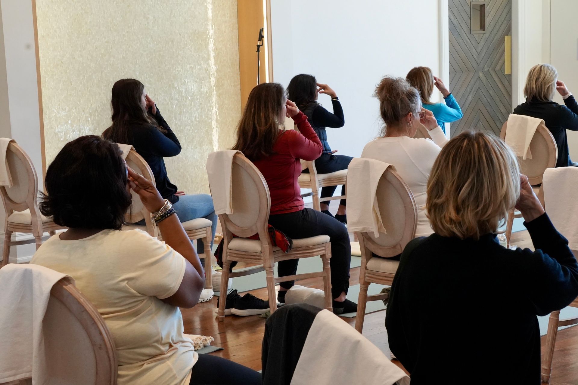 People in a brightly lit room doing seated exercises, hands on their heads. Chairs with white towels.