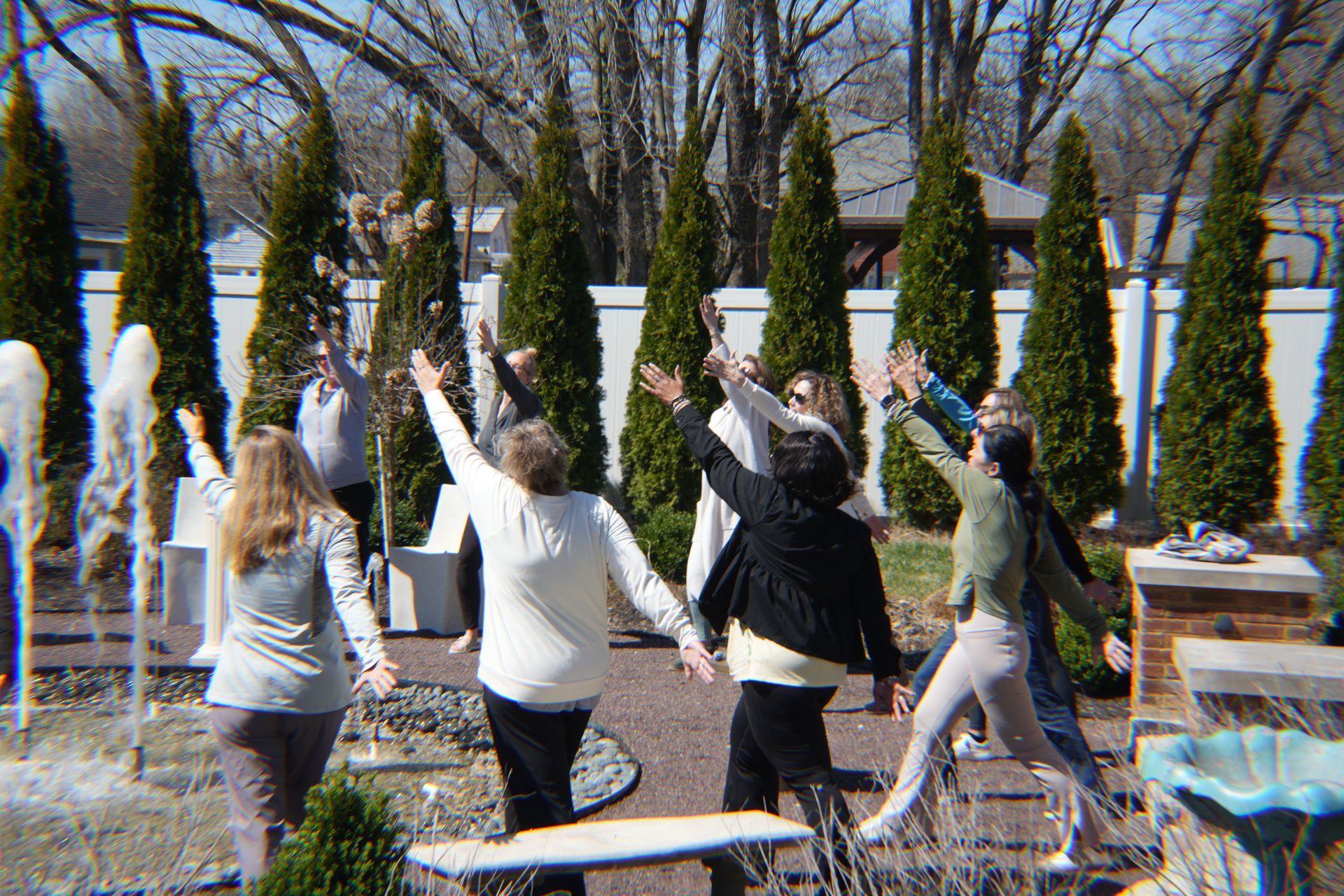 People with arms raised in a garden with fountains and tall green trees. Sunny day.