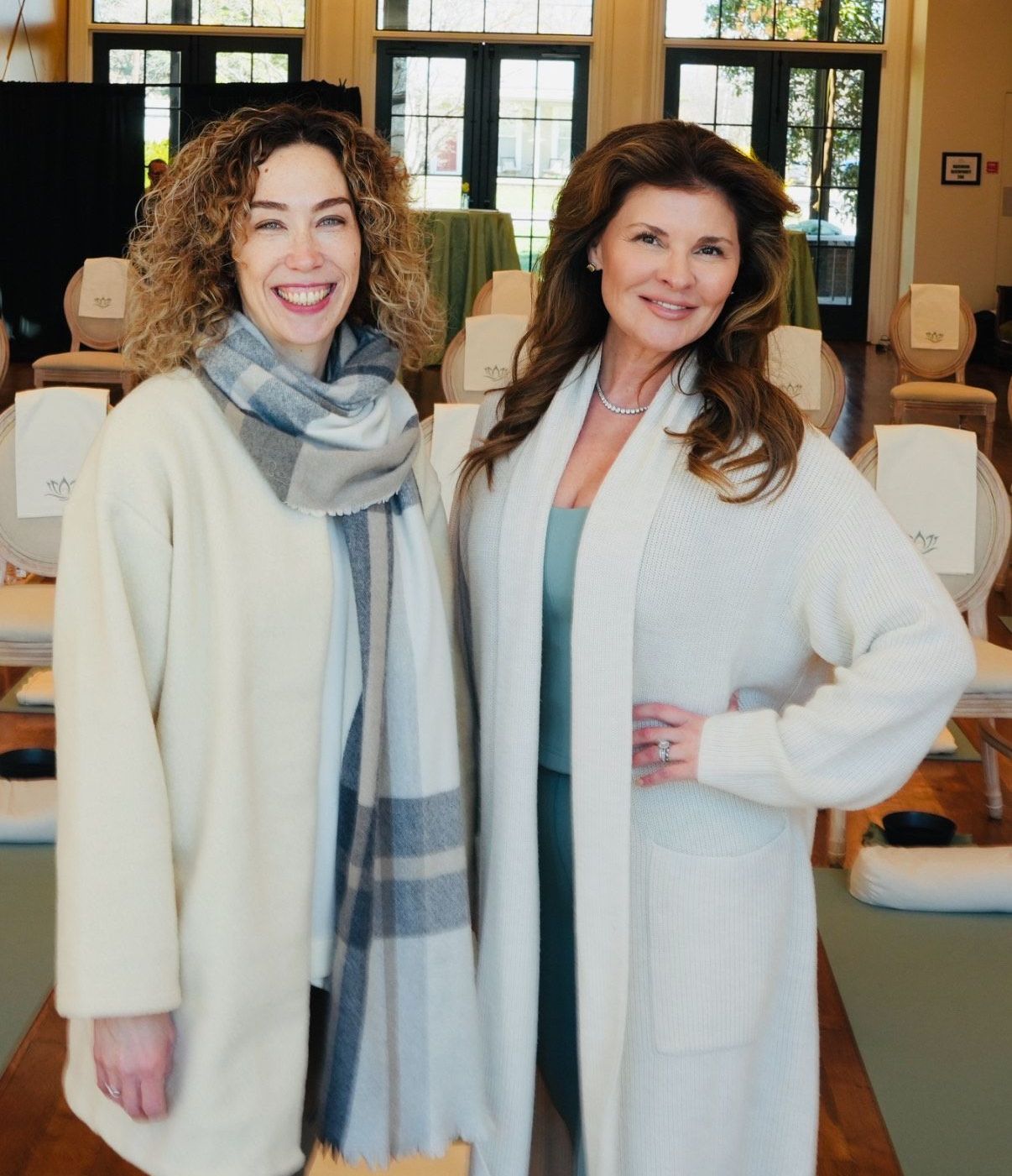 Two women pose together; one wears a cream coat and scarf, the other, a cardigan. They smile in a room with chairs.
