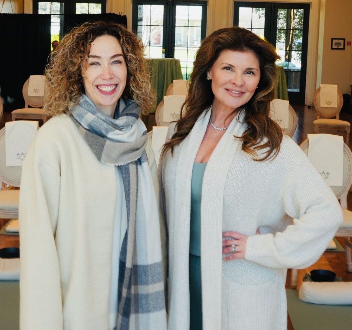 Two smiling women pose in a bright room with singing bowls. One wears a scarf, the other a cardigan.