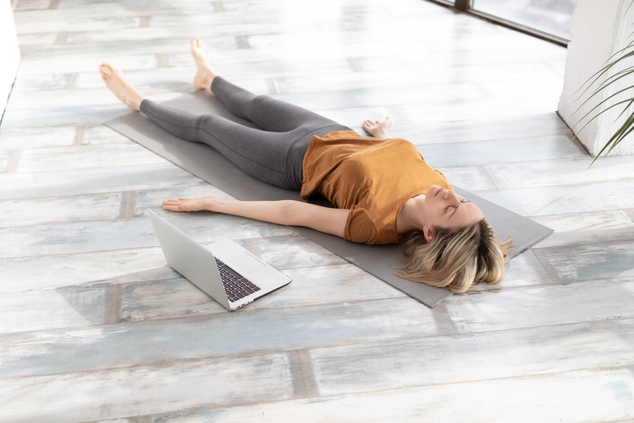 Woman in yoga pose on mat, laptop nearby. Indoors, natural light.