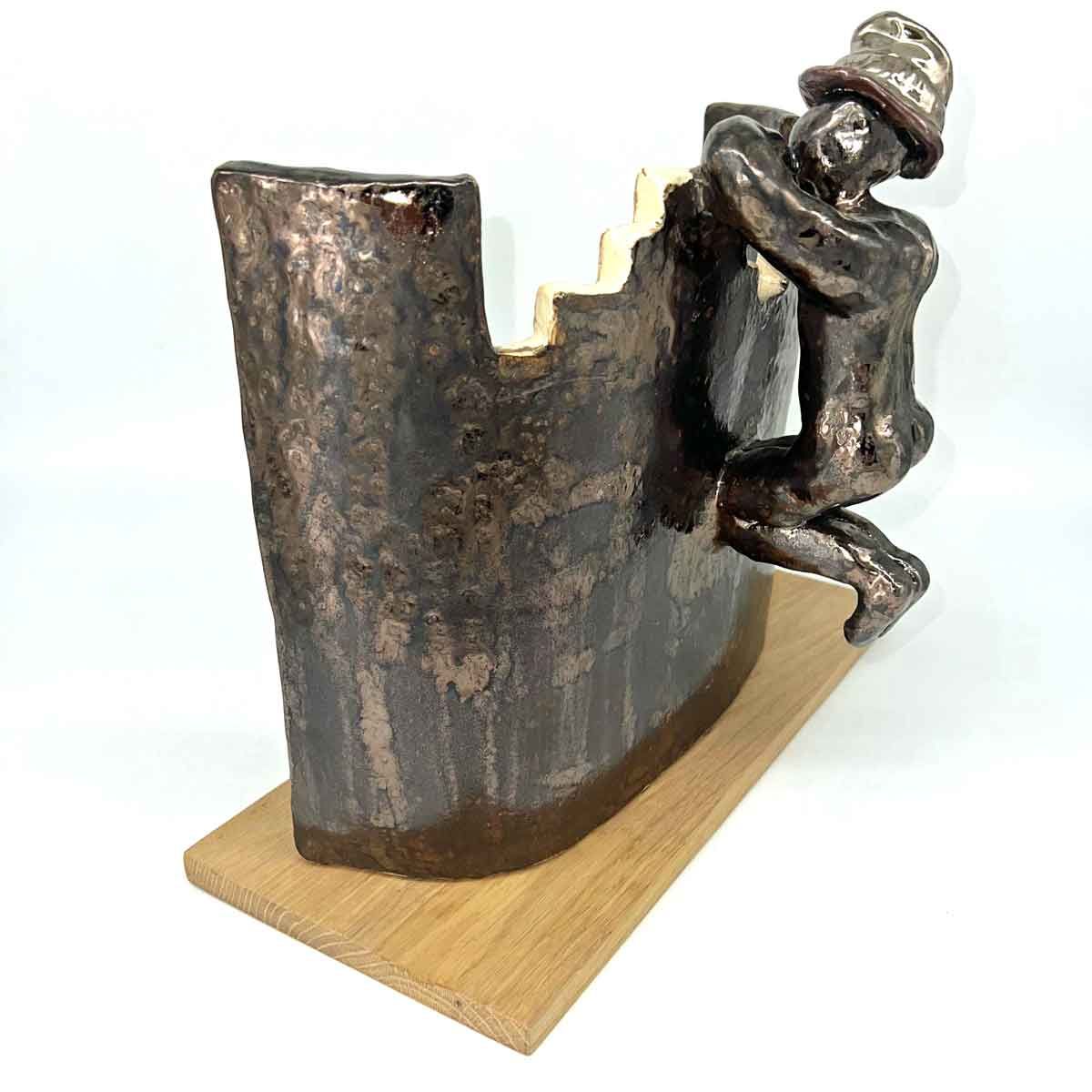 Klein bronzen beeldje dat tegen een donkere, gebogen metalen muur op een houten sokkel klimt.
