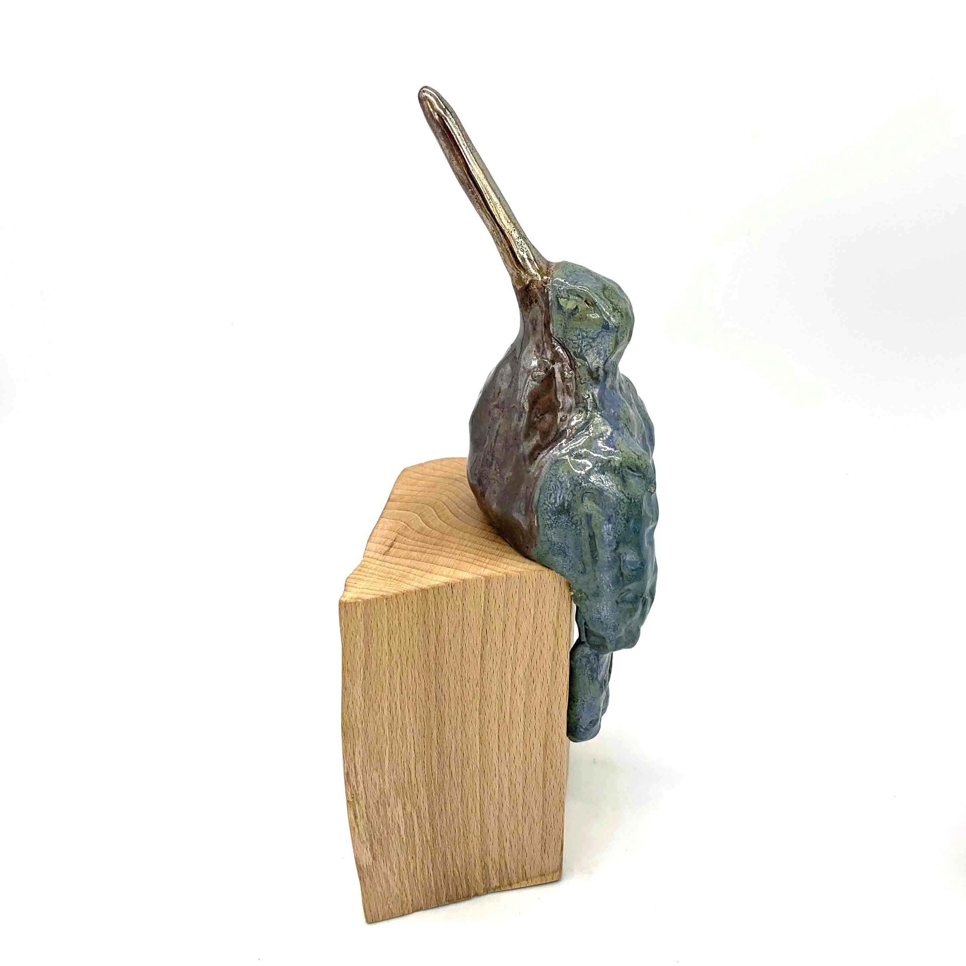 Klein blauwgroen vogelbeeldje, geplaatst op een houten blok tegen een witte achtergrond.