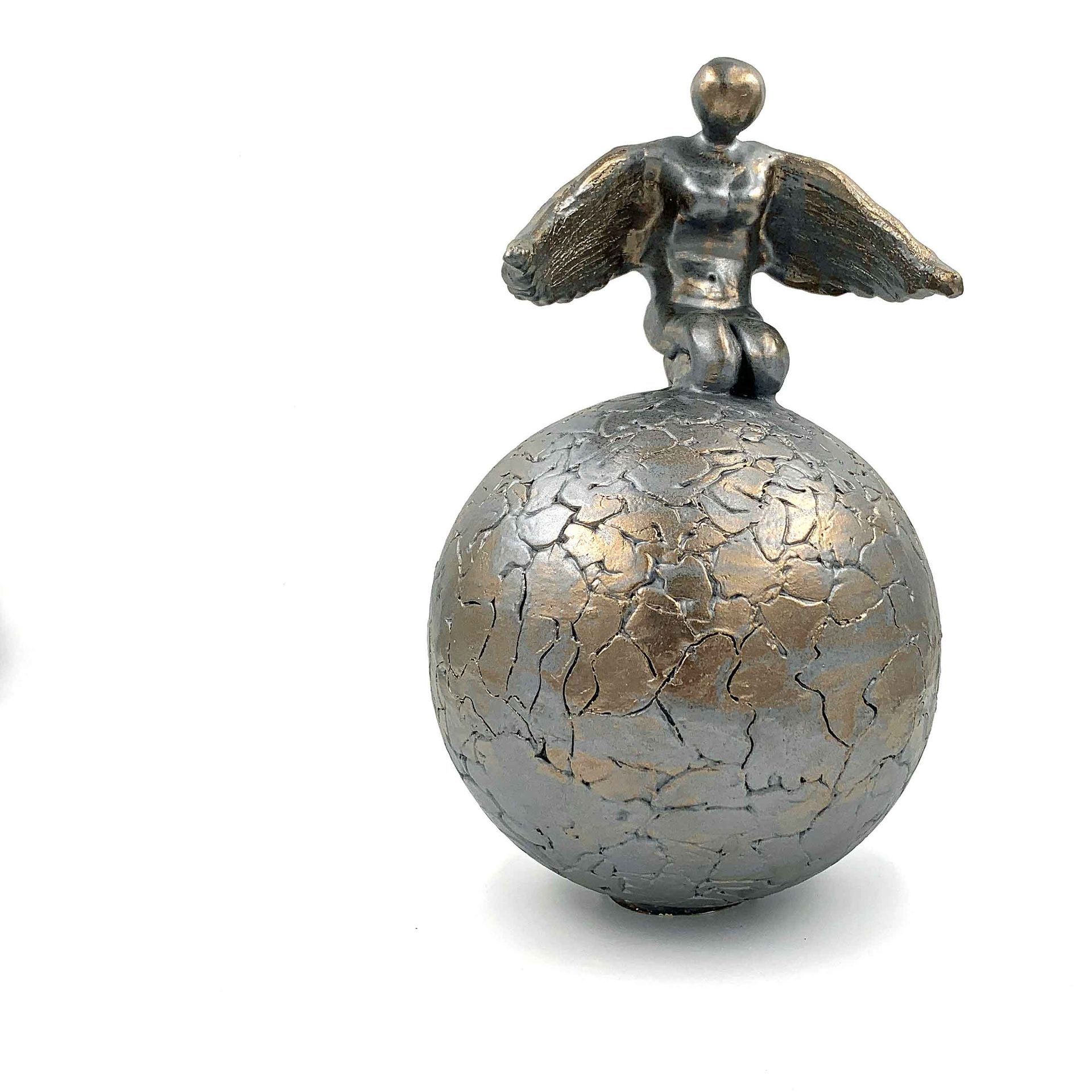Klein metalen beeldje met uitgestrekte armen, staand op een gebarsten zilveren bol.