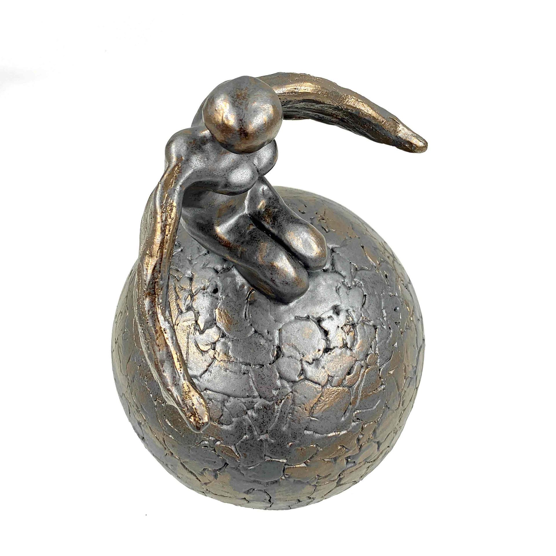 Klein metalen beeldje van een figuur zittend op een gebarsten zilveren bol met een gebogen arm of staart erboven.