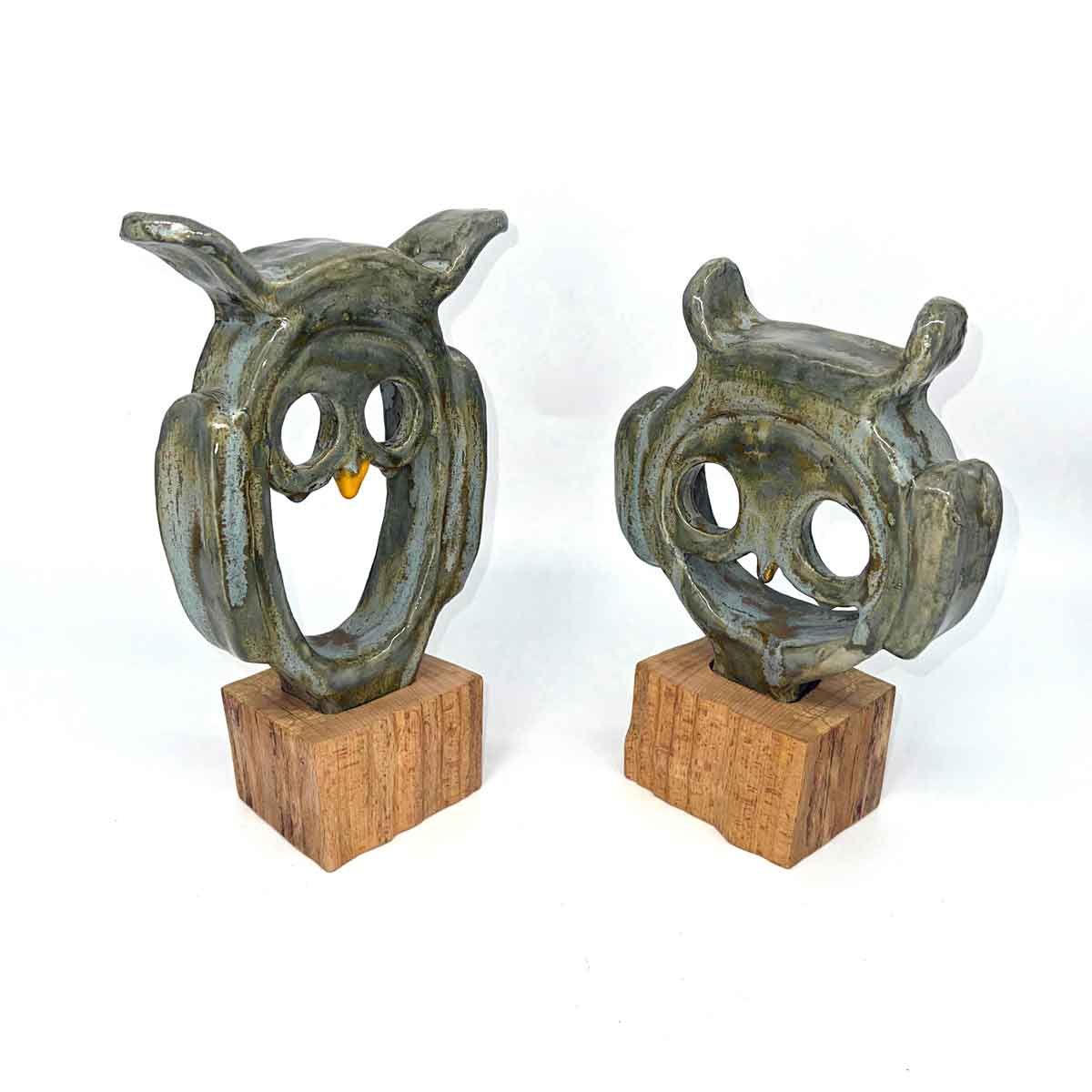 Twee kleine, groenachtige, uilachtige sculpturen op houten sokkels tegen een witte achtergrond.