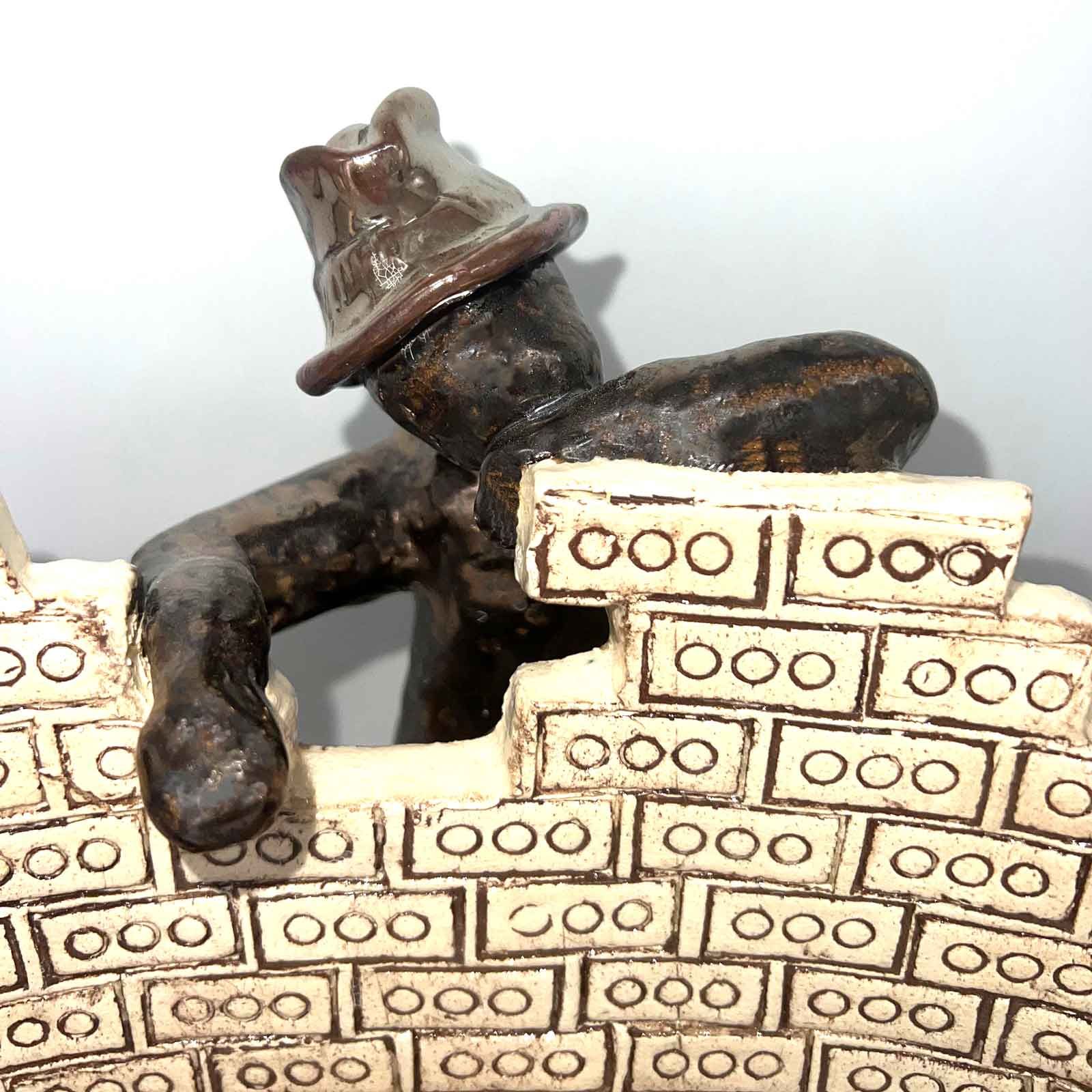 Een klein, donker figuurtje met een hoedje leunt over een bakstenen muur, gemaakt van speelgoedblokken.