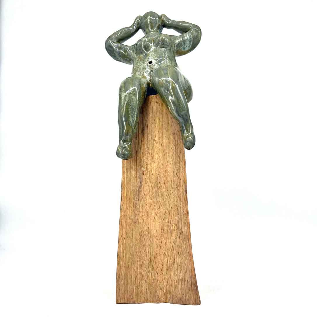 Klein groen beeldje zittend op een houten wig, met de handen naar het hoofd geheven.