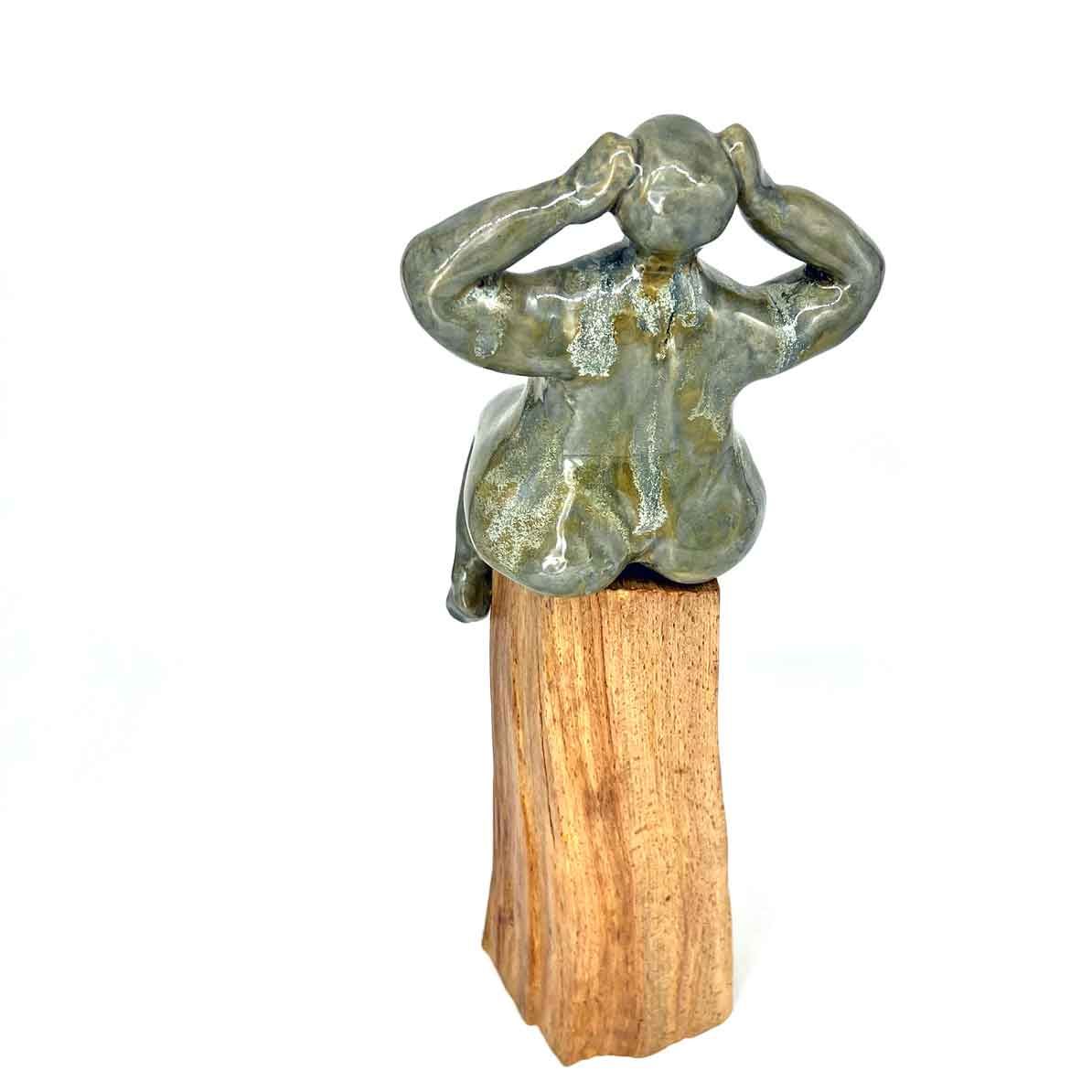 Klein metalen beeldje met handen op het hoofd, gemonteerd op een houten voetstuk tegen een witte achtergrond.