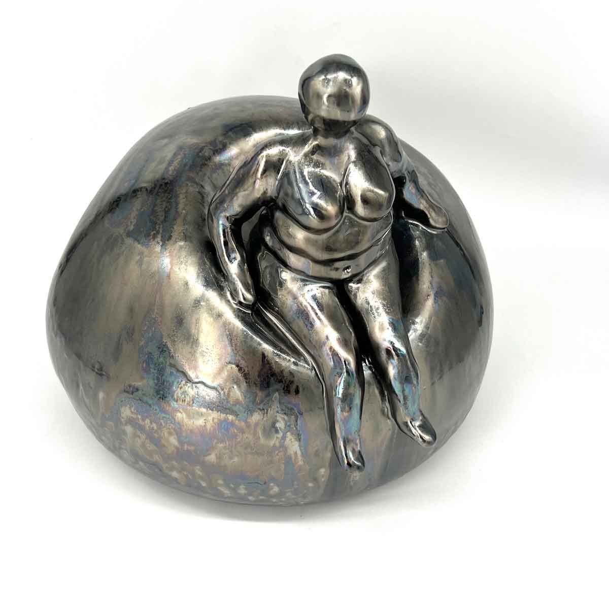 Metalen sculptuur van een zittende menselijke figuur met uitgestrekte benen op een afgerond zilveren oppervlak.
