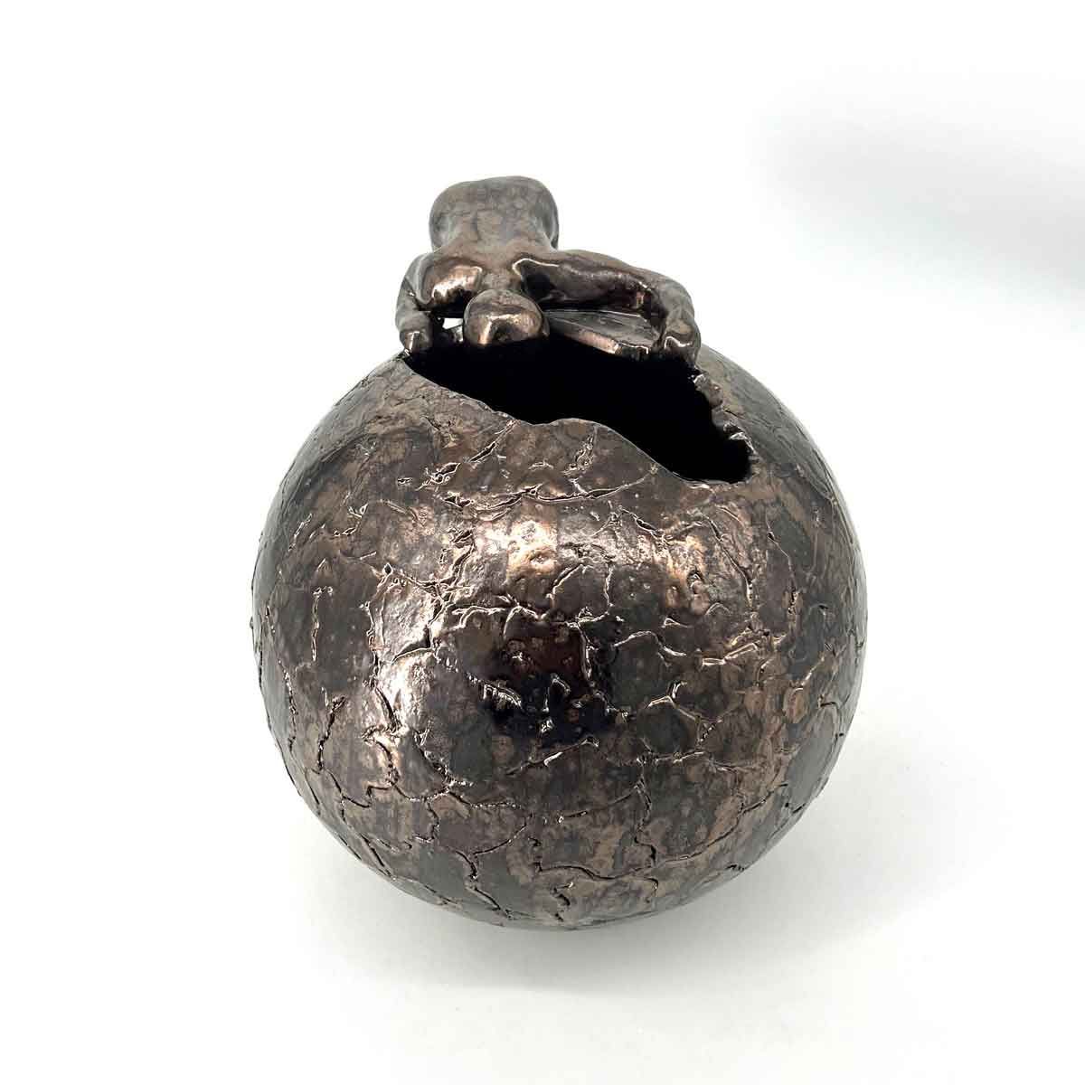 Een bolvormige metalen sculptuur met barstjes, met daarop een klein figuurtje, tegen een witte achtergrond.