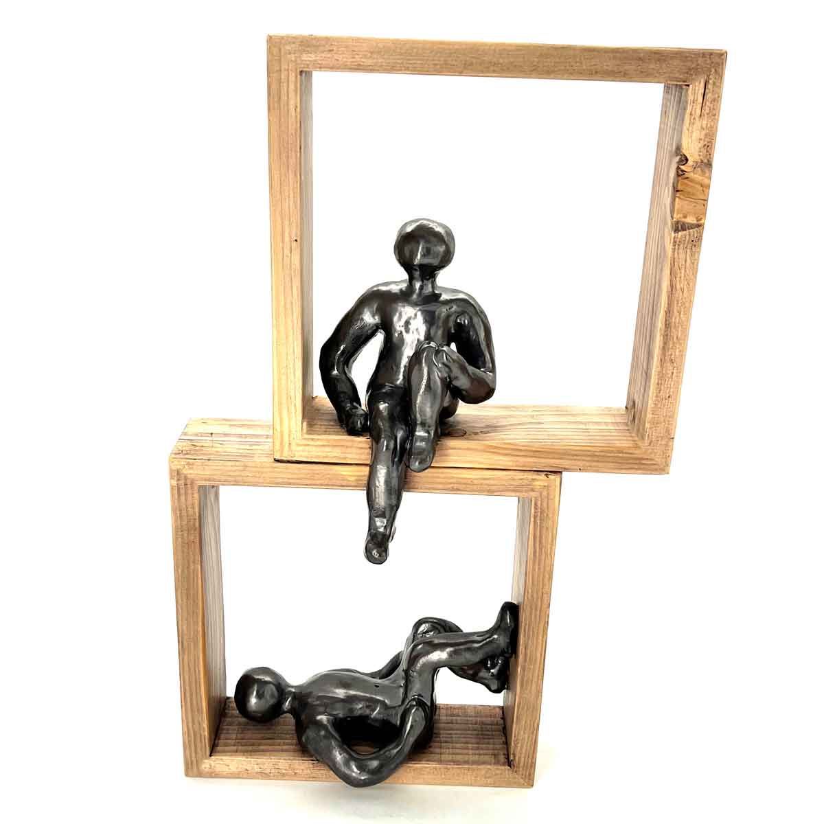 Twee zwarte beeldjes in op elkaar gestapelde vierkante houten frames, één zittend bovenop en één liggend eronder.