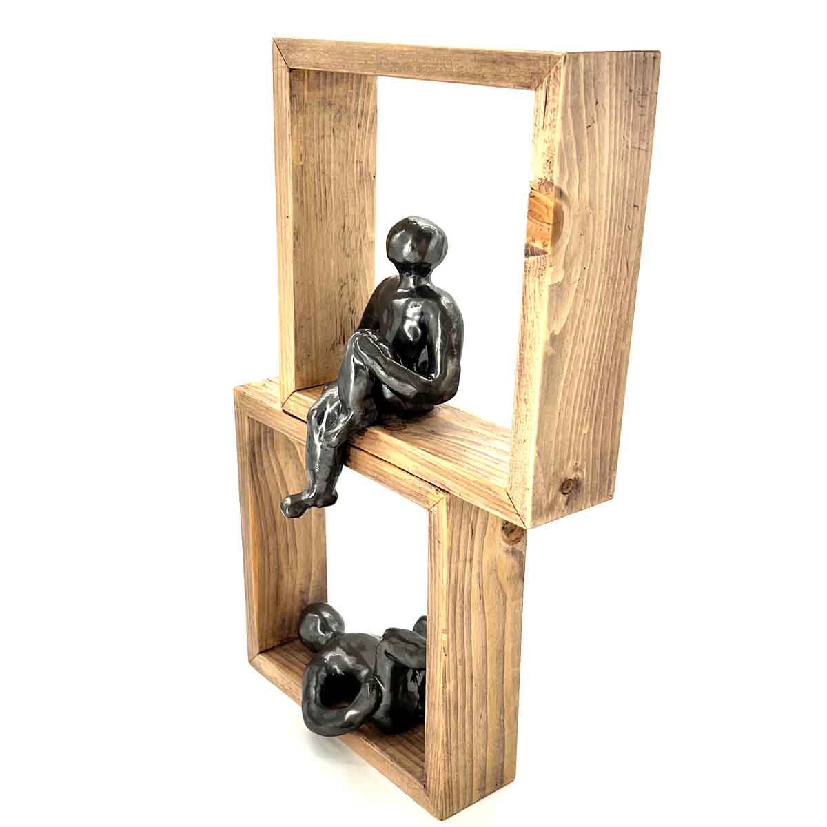 Zwarte beeldjes, zittend en liggend, op gestapelde houten kubusplanken in een minimalistische sculptuur.
