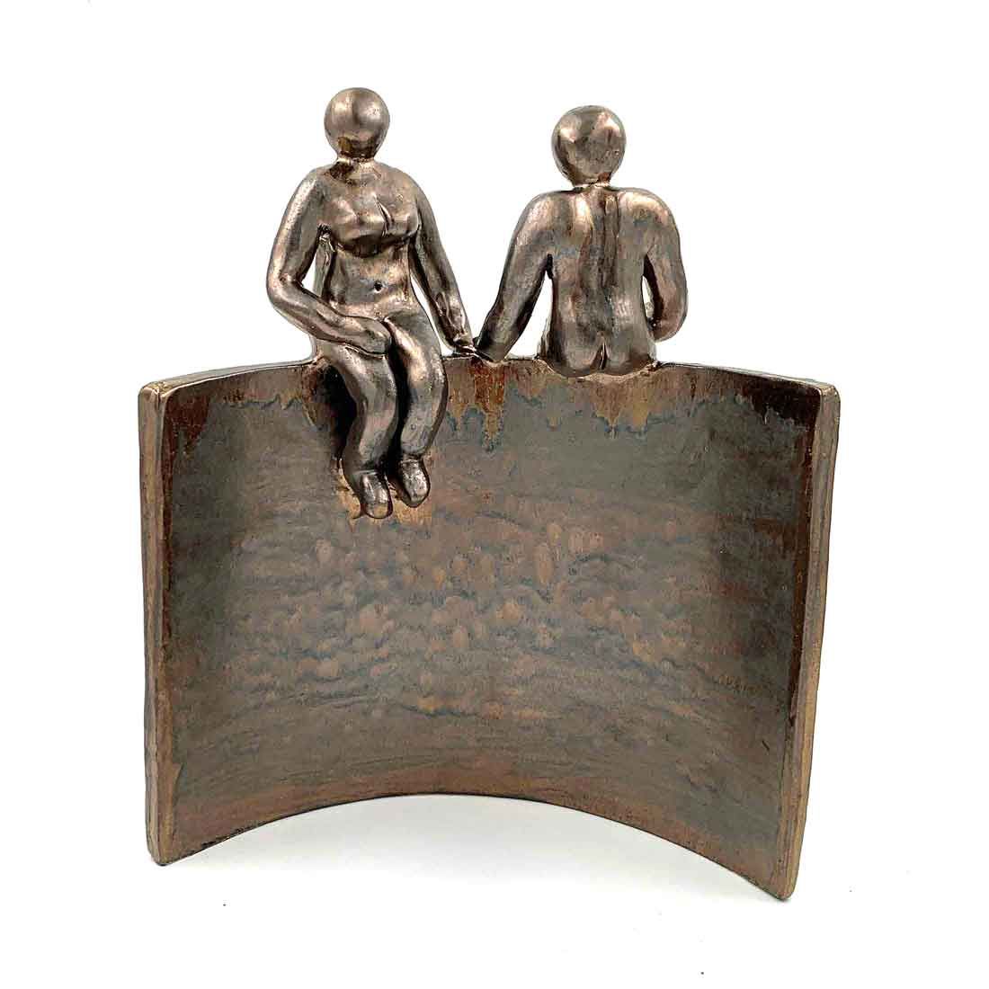 Twee kleine metalen figuurtjes zitten bovenop een gebogen bronzen plaat.