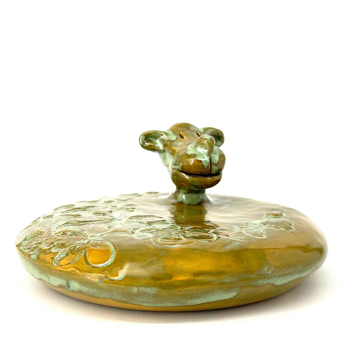 Een klein amberkleurig glazen schaaltje met een groenachtig deksel in de vorm van een dier, op een witte achtergrond.