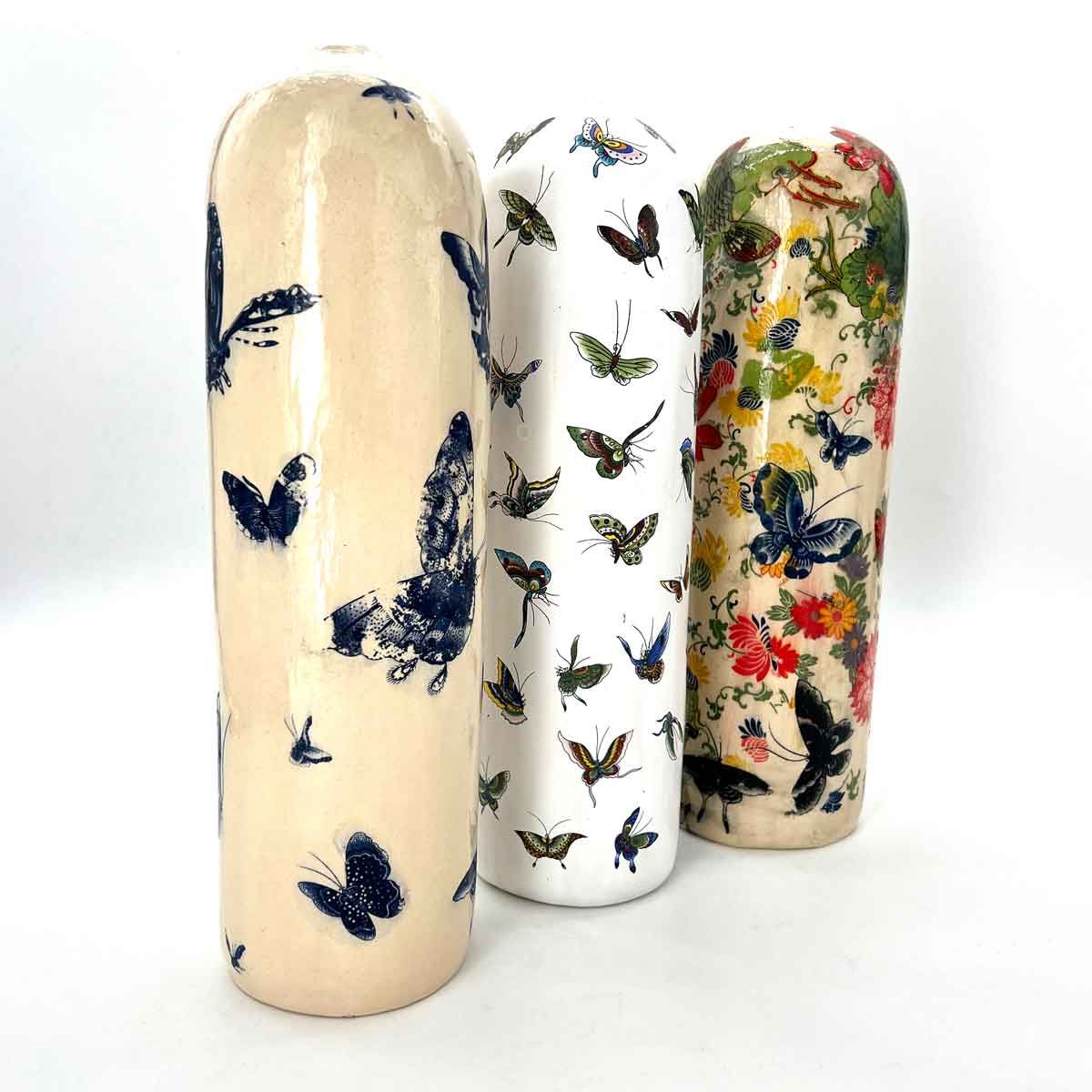 Drie decoratieve cilindrische vazen ​​met vlinderpatronen in crèmekleur, wit en bloemmotieven.